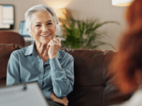 Older woman smiling during a conversation on a couch in a cozy living room setting.